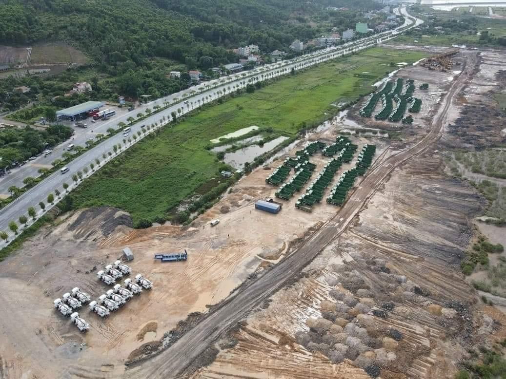 Toàn cảnh công trường Vinhomes Hạ Long Xanh với hàng loạt xe tải, máy xúc và thiết bị xây dựng trên khu đất rộng lớn, gần đường cao tốc và vùng đất ngập nước, với khung cảnh thiên nhiên xung quanh.
