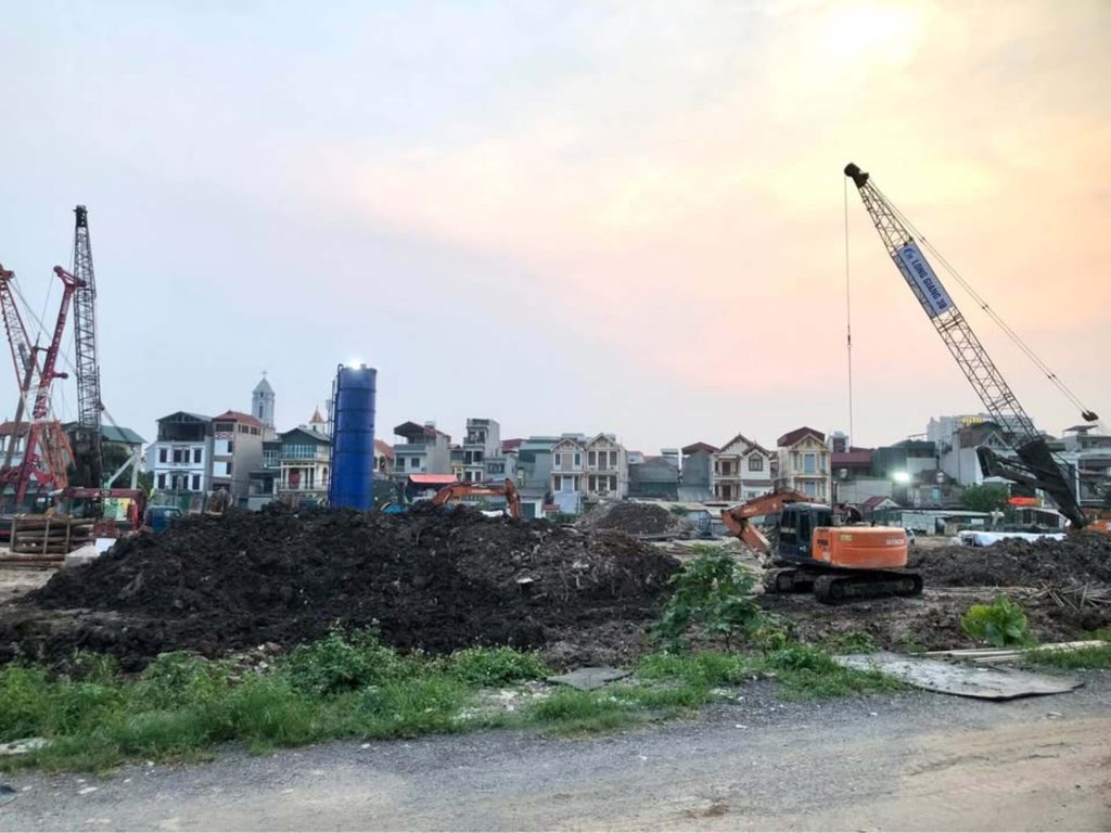 Galia Hanoi 19 Công trường dự án Galia Hanoi với máy xúc màu cam san đất, cần cẩu lớn vận chuyển vật liệu, và khu vực đất được đào xới vào buổi chiều, xung quanh là nhà dân và tòa nhà cao tầng.