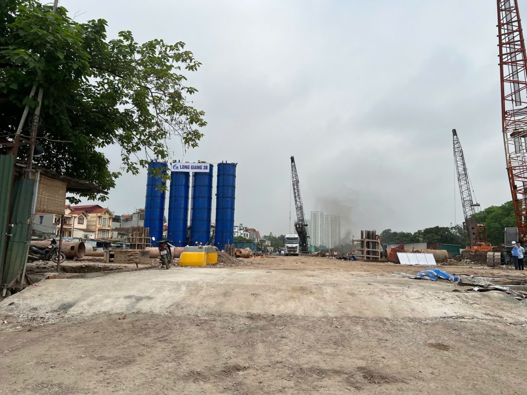 Galia Hanoi 18 Công trường dự án Galia Hanoi vào ban ngày, với tháp trộn bê tông màu xanh, cần cẩu lớn, và các thiết bị thi công móng, xung quanh là khu dân cư với nhà thấp tầng và cây xanh.