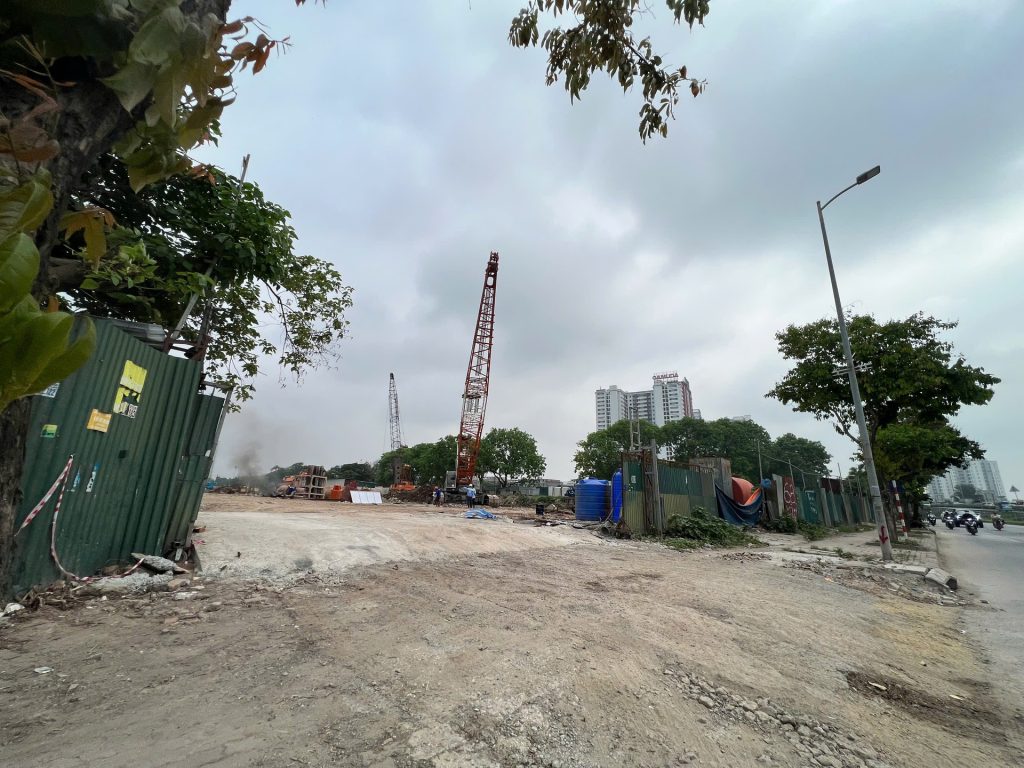Galia Hanoi 20 Công trường dự án Galia Hanoi với cần cẩu lớn màu đỏ, khu vực san nền bằng đất, hàng rào xanh bao quanh, và các tòa nhà cao tầng ở phía xa dưới bầu trời nhiều mây.